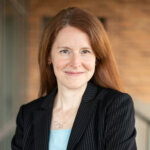 Woman with long red hair, black pinstripe blazer, smiling, standing with arms crossed.