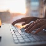Hands typing on a laptop with a sunset reflection on the screen.