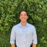 Smiling person in a light blue shirt stands against a lush green leafy background.