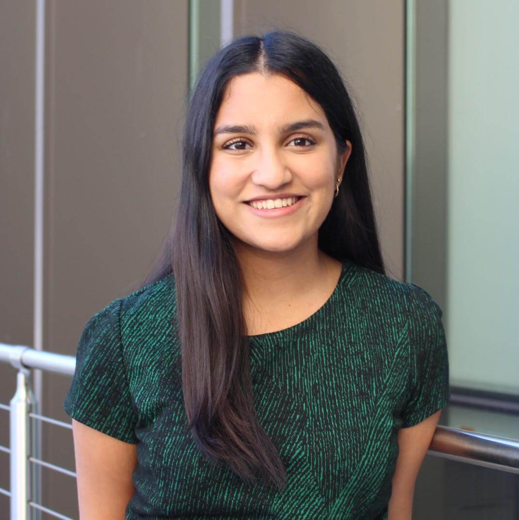 Smiling woman with long dark hair in a green patterned dress, standing by a railing.