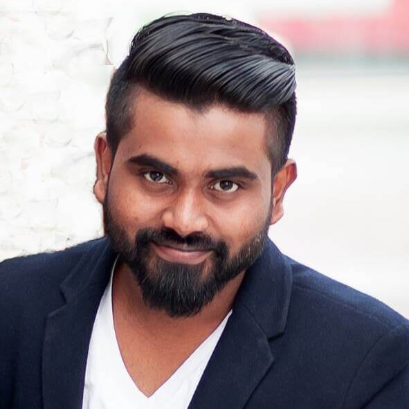 Man with neatly styled dark hair and beard, wearing a navy blazer over a white shirt, leaning against a reflective surface.