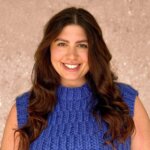 Smiling person with long brown hair wearing a blue knitted sleeveless top, against a textured beige background.