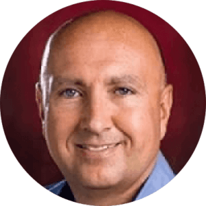 Smiling bald man against a dark red background, wearing a blue collared shirt.