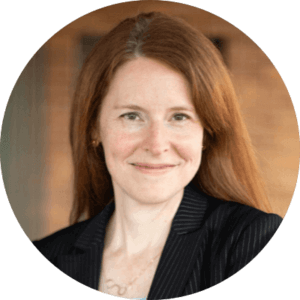 Smiling woman with long red hair wearing a dark pinstripe blazer, standing in front of a blurred brown background.