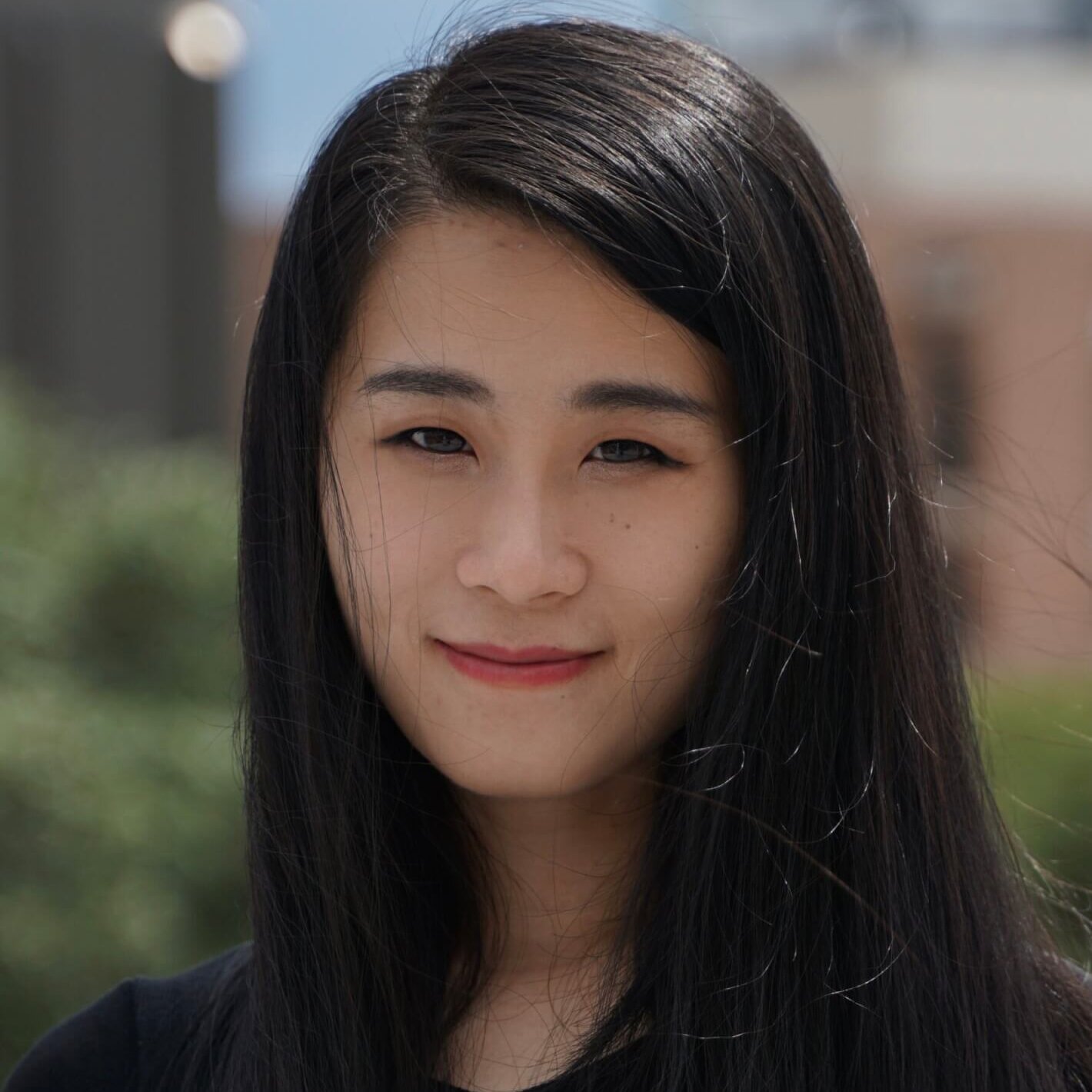 Woman with long black hair smiling softly, wearing a black shirt, standing outdoors with a blurred background.
