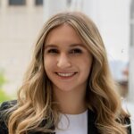 Smiling woman with long, wavy blonde hair in a formal outfit, standing outdoors.