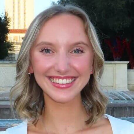 Smiling person with short blonde hair, outdoor background with trees and a building.